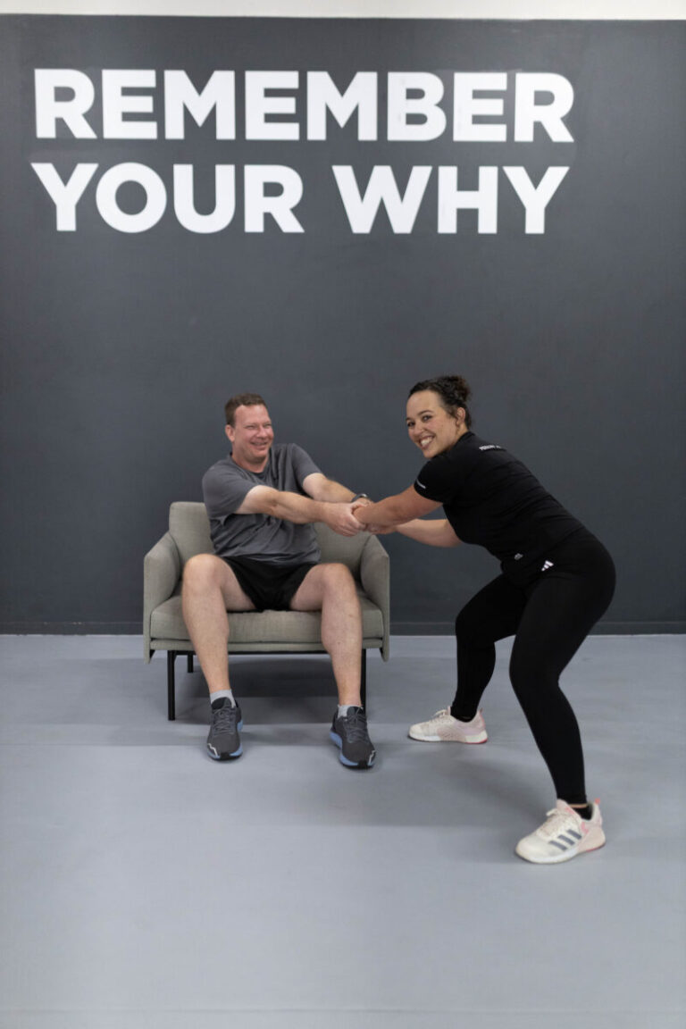 Marcia Swanepoel, a personal trainer at Virgin Active The Sanctuary, playfully dragging a client out of a chair to work out in a gym with 'YOUR WHY' on the wall, inspiring motivation with a fun approach.