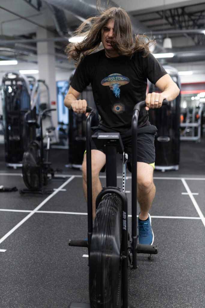 A client enjoying an energetic cardio session on an air bike at Virgin Active The Sanctuary in Somerset West, with flowing hair adding a playful vibe, emphasizing the fun of cardio while highlighting the importance of balancing effort to avoid overwork for sustained heart health.
