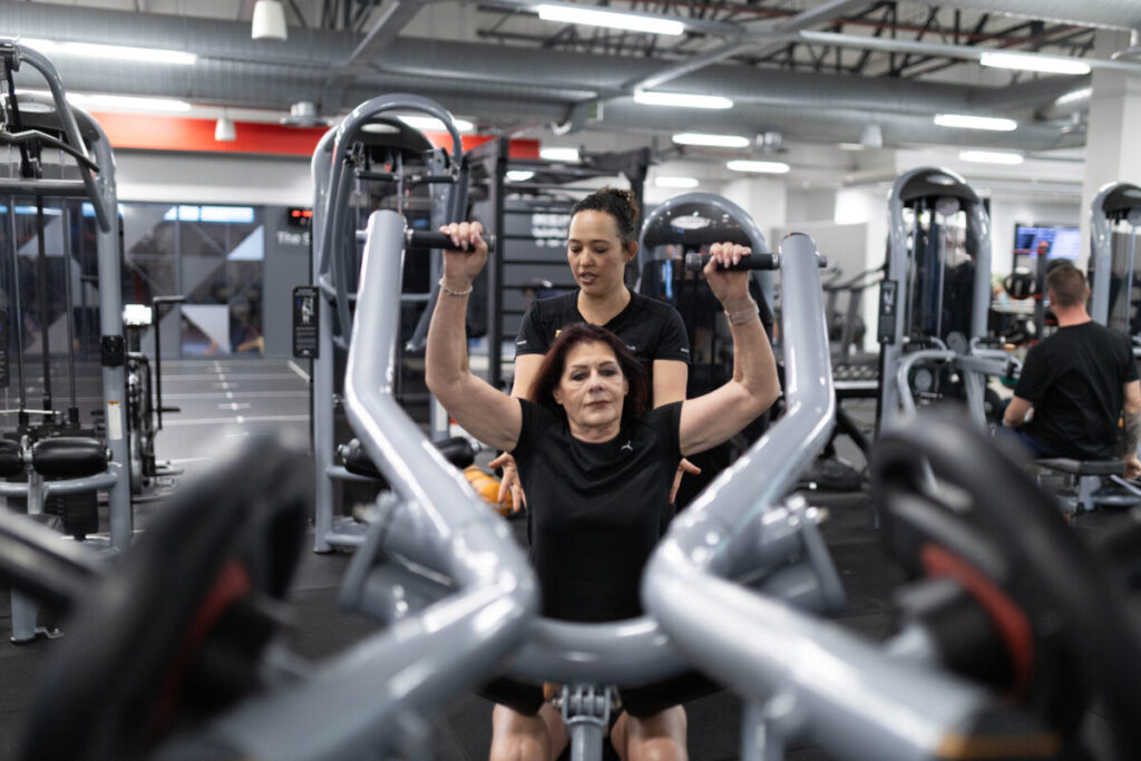 Marcia Swanepoel, a personal trainer at Virgin Active The Sanctuary in Somerset West, assisting a mature client with a strength exercise on a gym machine, promoting muscle health and fitness at any age.